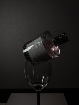 A dark, moody setting features a perfume bottle delicately balanced on the rim of a wine glass. The bottle is adorned with droplets of water, reflecting minimal light, creating a dramatic and elegant vibe. The background is a deep grey, adding to the sophisticated ambiance.