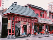 A vibrant scene from a street featuring a traditional architectural structure with a green-tiled roof and red accents. Nearby, a pink building houses a restaurant named Restoran Kiew Yee Baru, with signs in both English and Chinese. There is some activity with a person standing in front of the establishment, creating a lively atmosphere.