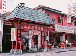 A vibrant scene from a street featuring a traditional architectural structure with a green-tiled roof and red accents. Nearby, a pink building houses a restaurant named Restoran Kiew Yee Baru, with signs in both English and Chinese. There is some activity with a person standing in front of the establishment, creating a lively atmosphere.