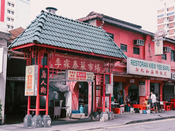 A vibrant scene from a street featuring a traditional architectural structure with a green-tiled roof and red accents. Nearby, a pink building houses a restaurant named Restoran Kiew Yee Baru, with signs in both English and Chinese. There is some activity with a person standing in front of the establishment, creating a lively atmosphere.