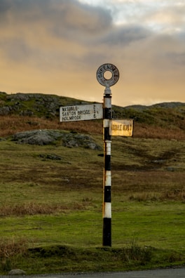 A vintage-style signpost stands in a rural landscape with hills in the background. The signpost is painted in black and white stripes with circular and rectangular signs indicating directions to nearby locations, including Wasdale and Greendale.