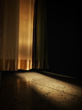 Curtains gently illuminated by sunlight, casting a warm glow on the floor. The light highlights the textured pattern of the fabric and creates a contrast between the bright and shadowed areas.