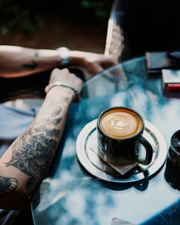 A tattooed arm rests on a glass table, next to a cup of coffee on a metal saucer. Sunlight creates natural patterns and reflections on the table surface, adding a tranquil ambiance. The coffee appears to be in a dark mug and has a latte art design on top.