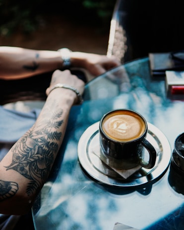 A tattooed arm rests on a glass table, next to a cup of coffee on a metal saucer. Sunlight creates natural patterns and reflections on the table surface, adding a tranquil ambiance. The coffee appears to be in a dark mug and has a latte art design on top.