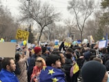 A large group of people gathered in a park, holding signs with messages such as 'Stop War' and displaying the European Union flag. Many individuals are wearing winter clothing. The background is filled with bare trees, and some people are holding balloons in blue and yellow, the colors of the Ukrainian flag.