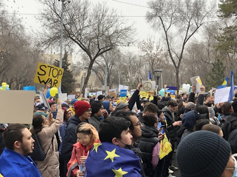 A large group of people gathered in a park, holding signs with messages such as 'Stop War' and displaying the European Union flag. Many individuals are wearing winter clothing. The background is filled with bare trees, and some people are holding balloons in blue and yellow, the colors of the Ukrainian flag.