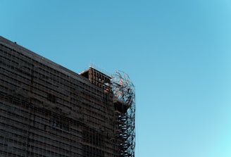 A building under construction with visible metal scaffolding on the side. The structure is partially wrapped in protective materials, and the sky is clear and blue.