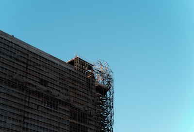 A building under construction with visible metal scaffolding on the side. The structure is partially wrapped in protective materials, and the sky is clear and blue.