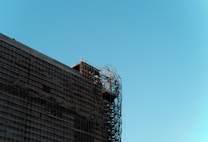 A building under construction with visible metal scaffolding on the side. The structure is partially wrapped in protective materials, and the sky is clear and blue.