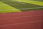 A sports field with vivid green grass divided into sections by white lines in the background. In the foreground, a track with a reddish-brown surface is marked with numbers and lines.