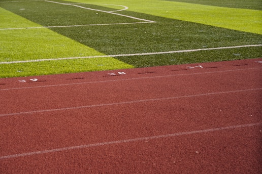 A sports field with vivid green grass divided into sections by white lines in the background. In the foreground, a track with a reddish-brown surface is marked with numbers and lines.