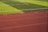 A sports field with vivid green grass divided into sections by white lines in the background. In the foreground, a track with a reddish-brown surface is marked with numbers and lines.