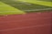 A sports field with vivid green grass divided into sections by white lines in the background. In the foreground, a track with a reddish-brown surface is marked with numbers and lines.