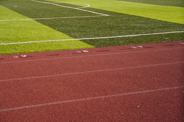 A sports field with vivid green grass divided into sections by white lines in the background. In the foreground, a track with a reddish-brown surface is marked with numbers and lines.