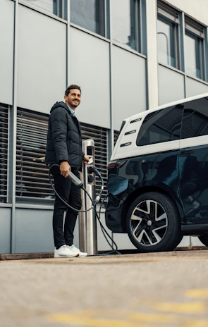 A person stands next to an electric vehicle that is connected to a charging station. The background consists of a modern building with large windows. The person is dressed in a dark jacket and white sneakers, holding the charging cable.