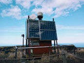 A solar-powered device is mounted on a platform in an outdoor setting, surrounded by a metal railing. Two large solar panels are prominently displayed on the front of the device, with a smaller white satellite dish and a dome-shaped object on top. The area around the platform is grassy with dry vegetation, and the background shows a vast expanse of blue sky with scattered clouds.
