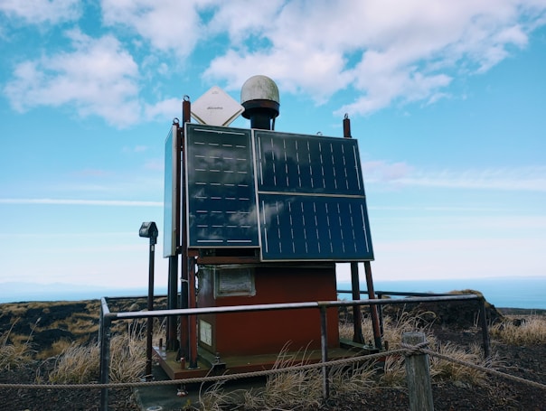 A solar-powered device is mounted on a platform in an outdoor setting, surrounded by a metal railing. Two large solar panels are prominently displayed on the front of the device, with a smaller white satellite dish and a dome-shaped object on top. The area around the platform is grassy with dry vegetation, and the background shows a vast expanse of blue sky with scattered clouds.