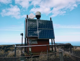 A solar-powered device is mounted on a platform in an outdoor setting, surrounded by a metal railing. Two large solar panels are prominently displayed on the front of the device, with a smaller white satellite dish and a dome-shaped object on top. The area around the platform is grassy with dry vegetation, and the background shows a vast expanse of blue sky with scattered clouds.
