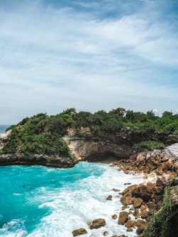 A rugged coastal landscape featuring turquoise waters crashing against rocky shores. Dense greenery covers the cliffs that surround a small cove, and a wooden path can be seen to the right side of the image.