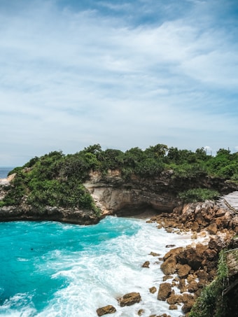 A rugged coastal landscape featuring turquoise waters crashing against rocky shores. Dense greenery covers the cliffs that surround a small cove, and a wooden path can be seen to the right side of the image.