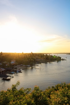 The image captures a serene waterfront scene at sunset, with a glowing sky transitioning from bright yellow near the horizon to vibrant blue above. An assortment of small boats is moored near wooden piers along the shoreline, lined with lush greenery and tropical palm trees. A few small buildings are nestled among the trees on the left side of the image.