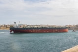 A large tanker ship is moving through a body of water against the backdrop of a cityscape. The ship has a red and black hull with white structures on the deck. The sky is partly cloudy, and the city in the background is composed of historic buildings with a uniform color scheme of beige and light brown. There is a tugboat assisting the tanker on the right side of the image.
