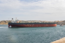 A large tanker ship is moving through a body of water against the backdrop of a cityscape. The ship has a red and black hull with white structures on the deck. The sky is partly cloudy, and the city in the background is composed of historic buildings with a uniform color scheme of beige and light brown. There is a tugboat assisting the tanker on the right side of the image.