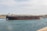 A large tanker ship is moving through a body of water against the backdrop of a cityscape. The ship has a red and black hull with white structures on the deck. The sky is partly cloudy, and the city in the background is composed of historic buildings with a uniform color scheme of beige and light brown. There is a tugboat assisting the tanker on the right side of the image.