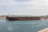 A large tanker ship is moving through a body of water against the backdrop of a cityscape. The ship has a red and black hull with white structures on the deck. The sky is partly cloudy, and the city in the background is composed of historic buildings with a uniform color scheme of beige and light brown. There is a tugboat assisting the tanker on the right side of the image.