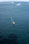 A small boat is visible in the foreground, navigating through calm, dark blue waters. In the distance, a larger vessel creates a wake as it moves toward a long pier with a lighthouse at the end. The sky is clear, and the overall scene suggests a tranquil maritime environment.