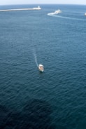 A small boat is visible in the foreground, navigating through calm, dark blue waters. In the distance, a larger vessel creates a wake as it moves toward a long pier with a lighthouse at the end. The sky is clear, and the overall scene suggests a tranquil maritime environment.