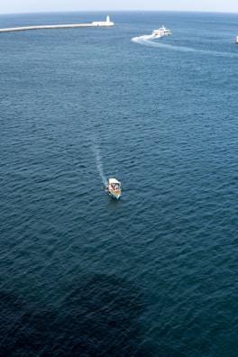 A small boat is visible in the foreground, navigating through calm, dark blue waters. In the distance, a larger vessel creates a wake as it moves toward a long pier with a lighthouse at the end. The sky is clear, and the overall scene suggests a tranquil maritime environment.