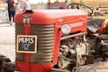 A vintage red tractor with a worn seat is parked outdoors, displaying a small chalkboard sign with the text 'MUMS $12.00'. In the background, people are visible at what appears to be a market or fair setting, along with additional flowers and plants.