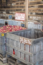 A rustic market setting with large wooden crates full of red apples. Various signs indicate instructions like 'Bag your own apples' and 'Fresh from the orchard. Please wash at home.' The scene portrays an inviting rural atmosphere with emphasis on fresh produce.