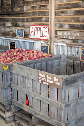 A rustic market setting with large wooden crates full of red apples. Various signs indicate instructions like 'Bag your own apples' and 'Fresh from the orchard. Please wash at home.' The scene portrays an inviting rural atmosphere with emphasis on fresh produce.