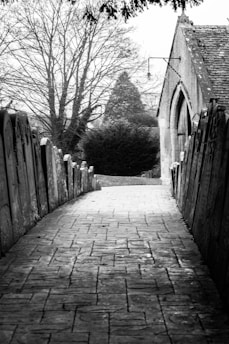 A black and white image of a narrow, cobblestone path flanked by a row of old tombstones on each side. In the background, there is a small stone building with an arched wooden door, and bare trees casting shadows over the scene.
