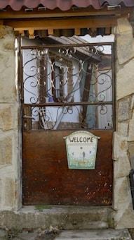 A rustic wooden and wrought iron gate is set within a stone frame. The gate features intricate swirling metal designs and a small welcome sign with decorative illustrations. Behind the gate, a quaint, weathered house with visible wooden beams and a partially tiled roof can be seen.