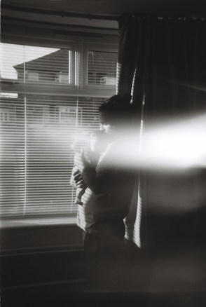 A person is holding a baby near a window with blinds partially open. The image is black and white with a soft light streak cutting across the frame. The scene appears intimate and serene, with the room dimly lit by natural light coming through the window.