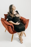 A pregnant woman in a black dress is seated comfortably in a red armchair. Her hands rest gently on her stomach, and her expression appears peaceful and serene. She wears white high-heeled sandals and her dark hair falls loosely over her shoulders.