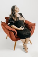 A pregnant woman in a black dress is seated comfortably in a red armchair. Her hands rest gently on her stomach, and her expression appears peaceful and serene. She wears white high-heeled sandals and her dark hair falls loosely over her shoulders.