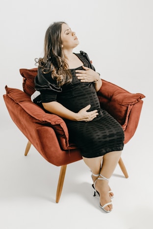 A pregnant woman in a black dress is seated comfortably in a red armchair. Her hands rest gently on her stomach, and her expression appears peaceful and serene. She wears white high-heeled sandals and her dark hair falls loosely over her shoulders.