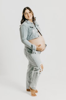A smiling pregnant woman is standing barefoot in a light denim outfit, consisting of a cropped jacket and ripped jeans. She is displaying her baby bump with both hands gently resting on it. The background is plain and white, emphasizing the subject.