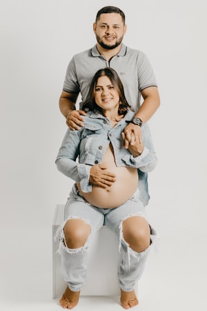 A couple is posing together, with a woman seated and a man standing behind her. The woman is pregnant, wearing a light denim jacket and ripped jeans, and seated barefoot on a white block or chair. The man is wearing a grey polo shirt and jeans, gently holding the woman's shoulders and her hand.