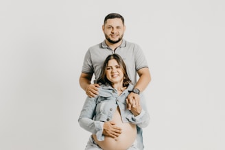 A couple is posed against a plain light gray background. The man stands behind the woman, placing his hands on her shoulders. The woman is sitting, smiling, and wearing a denim jacket open to reveal her pregnant belly.