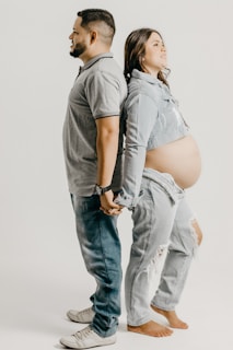A couple stands back-to-back holding hands. The man is wearing a gray polo shirt and jeans, while the woman is wearing a denim jacket over a bare belly, suggesting pregnancy, and ripped jeans. Both appear to be relaxed and content.