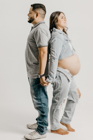 A couple stands back-to-back holding hands. The man is wearing a gray polo shirt and jeans, while the woman is wearing a denim jacket over a bare belly, suggesting pregnancy, and ripped jeans. Both appear to be relaxed and content.