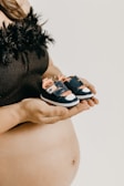 A person is holding a pair of small baby shoes in their hands, with a focus on the bare midriff showcasing a pregnant belly. The shoes are blue with pink and white accents, and the person is wearing a black top with a fluffy texture at the top edge.