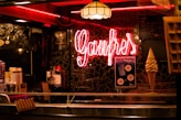 A warmly lit interior featuring a neon sign that reads 'Gaufres'. The setting includes a display of waffles and an ice cream cone. There is a menu written on a blackboard in the background alongside posters. The ambiance is cozy with a vintage feel, accented by a patterned wall and hanging lamps.