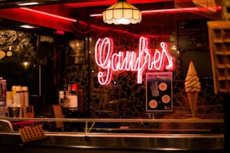 A warmly lit interior featuring a neon sign that reads 'Gaufres'. The setting includes a display of waffles and an ice cream cone. There is a menu written on a blackboard in the background alongside posters. The ambiance is cozy with a vintage feel, accented by a patterned wall and hanging lamps.