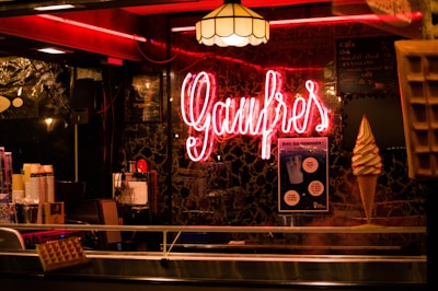 A warmly lit interior featuring a neon sign that reads 'Gaufres'. The setting includes a display of waffles and an ice cream cone. There is a menu written on a blackboard in the background alongside posters. The ambiance is cozy with a vintage feel, accented by a patterned wall and hanging lamps.